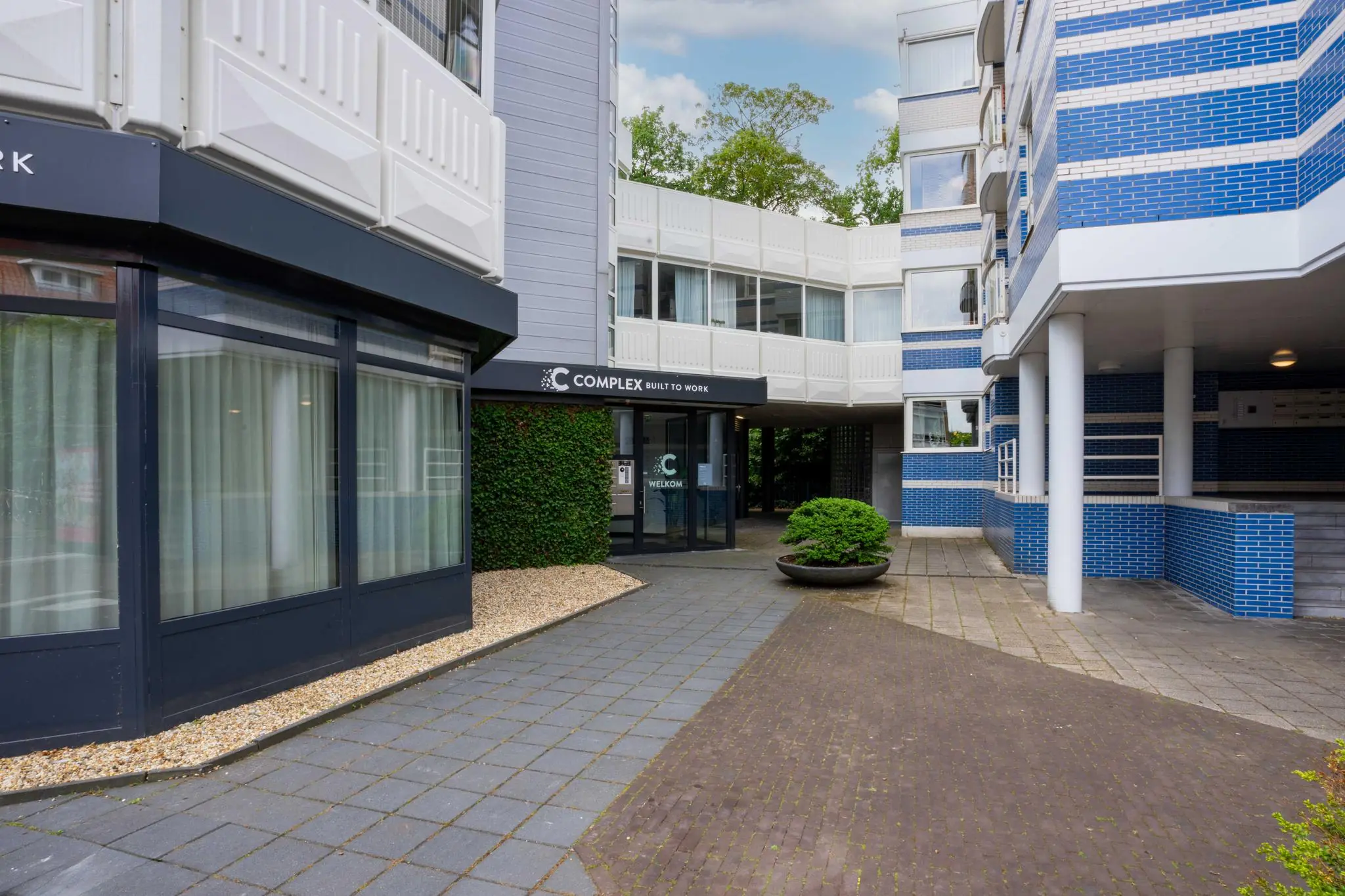 Ingang van het kantoorgebouw 'Complex Built to Work' aan de 1e Hogeweg, met moderne gevel en betegelde stoep.